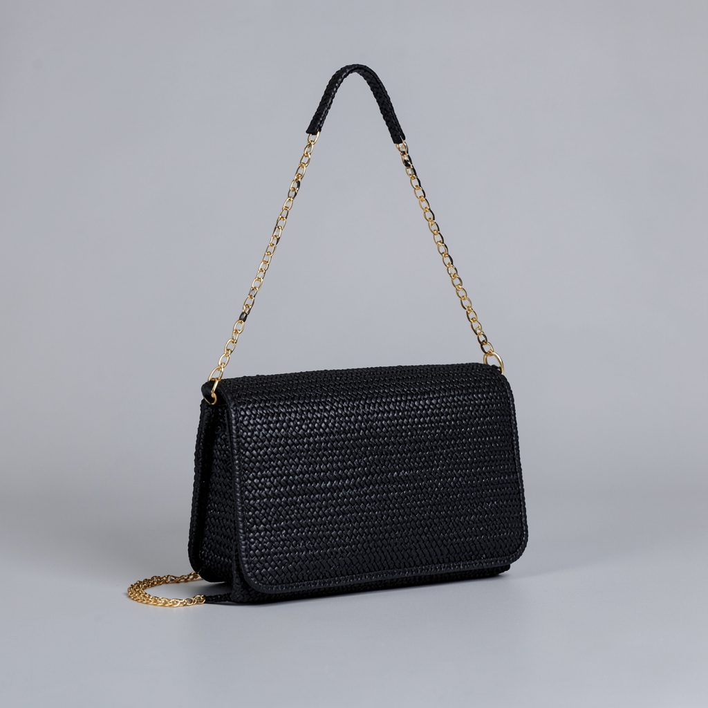 A black woven crossbody bag with an intricate textured pattern and a slim gold chain strap, set against a soft gray background