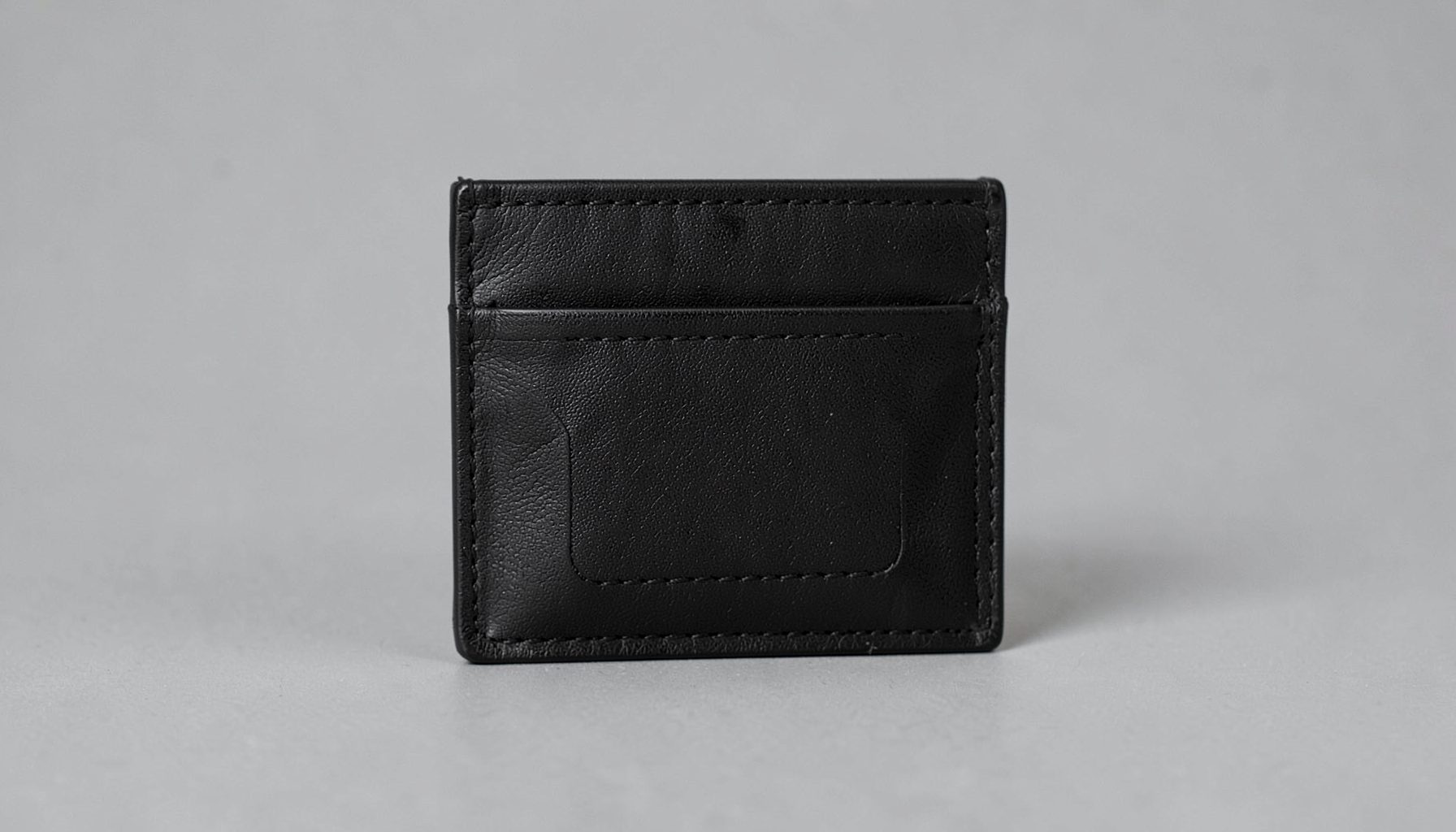 A compact black vegan leather cardholder with a central pocket, shown against a soft gray backdrop.