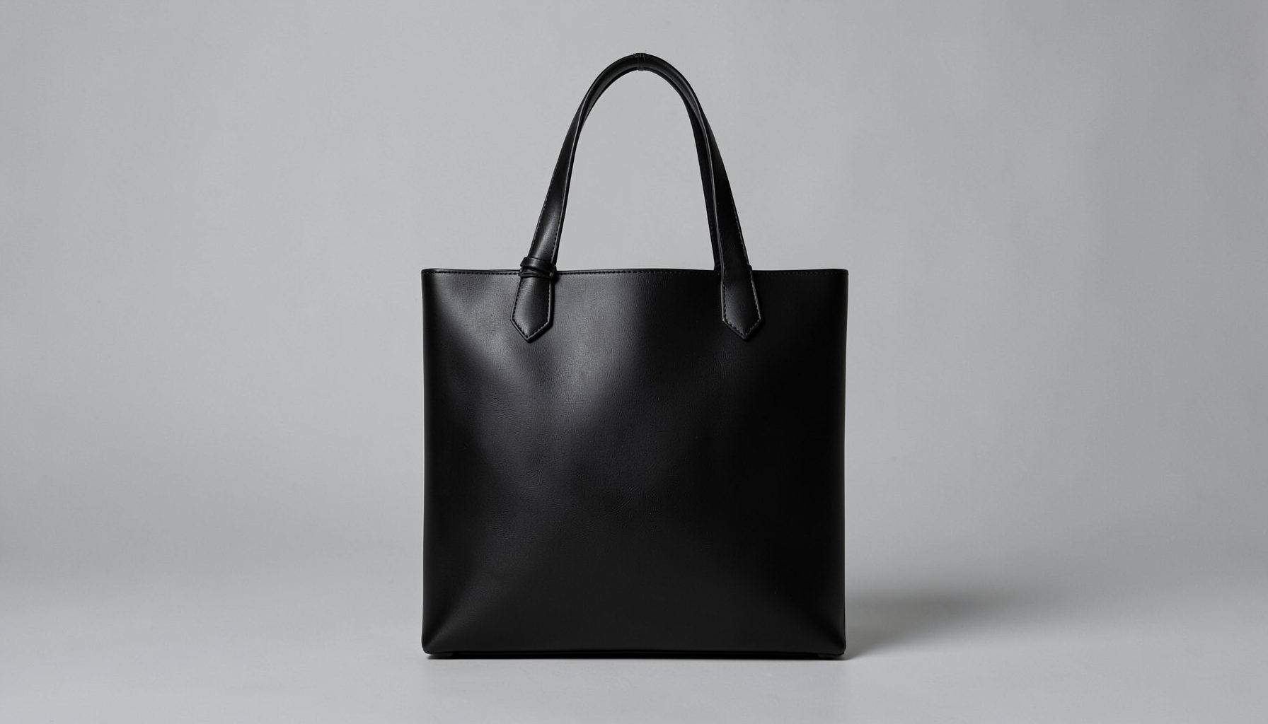 A sleek black vegan leather tote bag with clean lines, photographed against a soft gray background