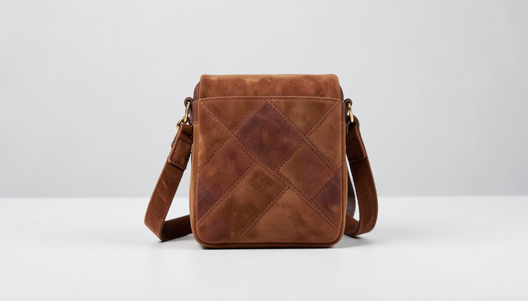 A sleek suede crossbody bag in a rich tone with geometric paneling and a removable strap, set against a minimalist backdrop.