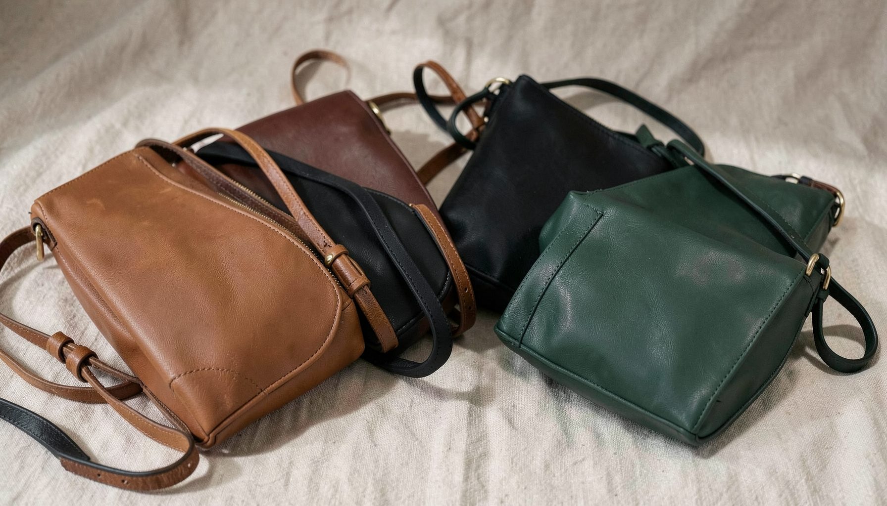 A softly lit arrangement of vegan leather crossbody bags in warm brown, black, and deep green tones, resting on a neutral linen surface with gentle shadows