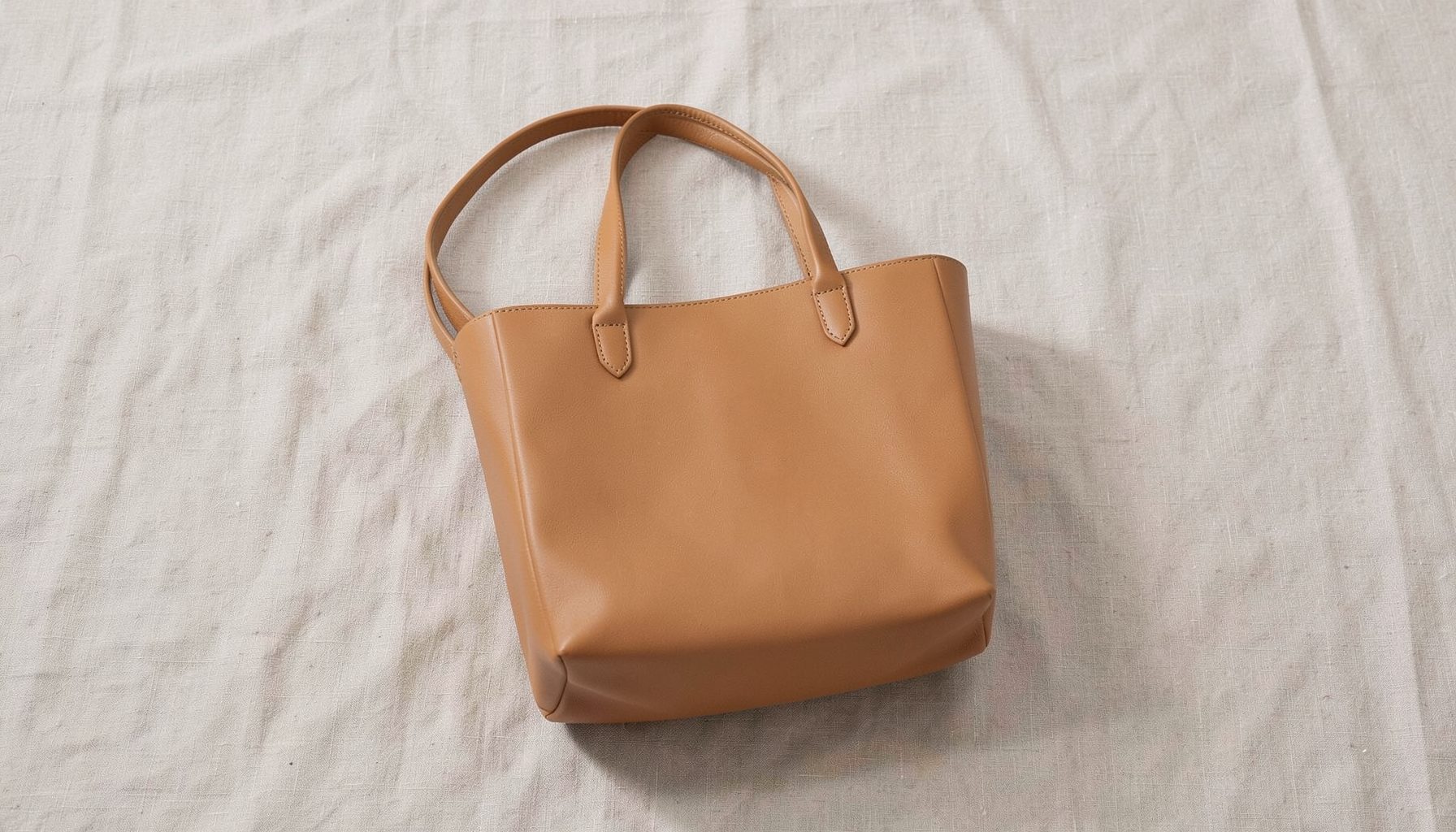 A softly lit flat lay of a structured vegan leather tote bag in warm tan, resting on a neutral linen surface with minimalist styling