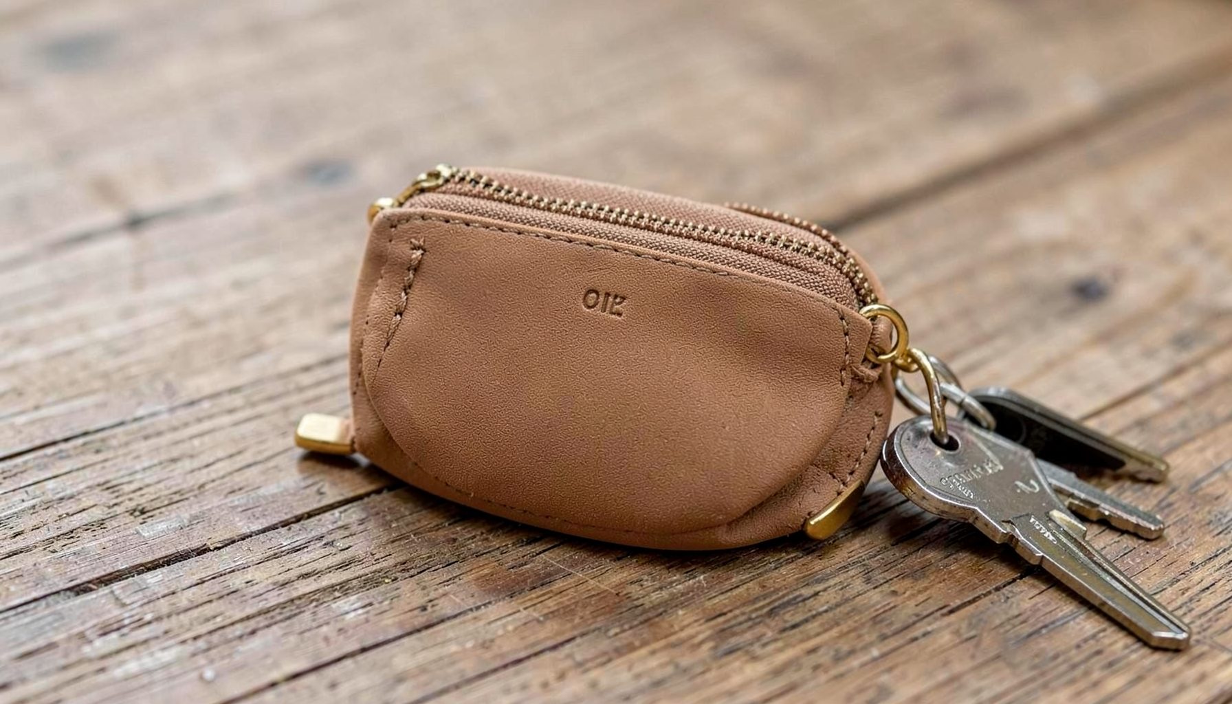 A zippered coin pouch in tan vegan leather with gold-toned hardware, resting beside a set of keys on a wooden table.
