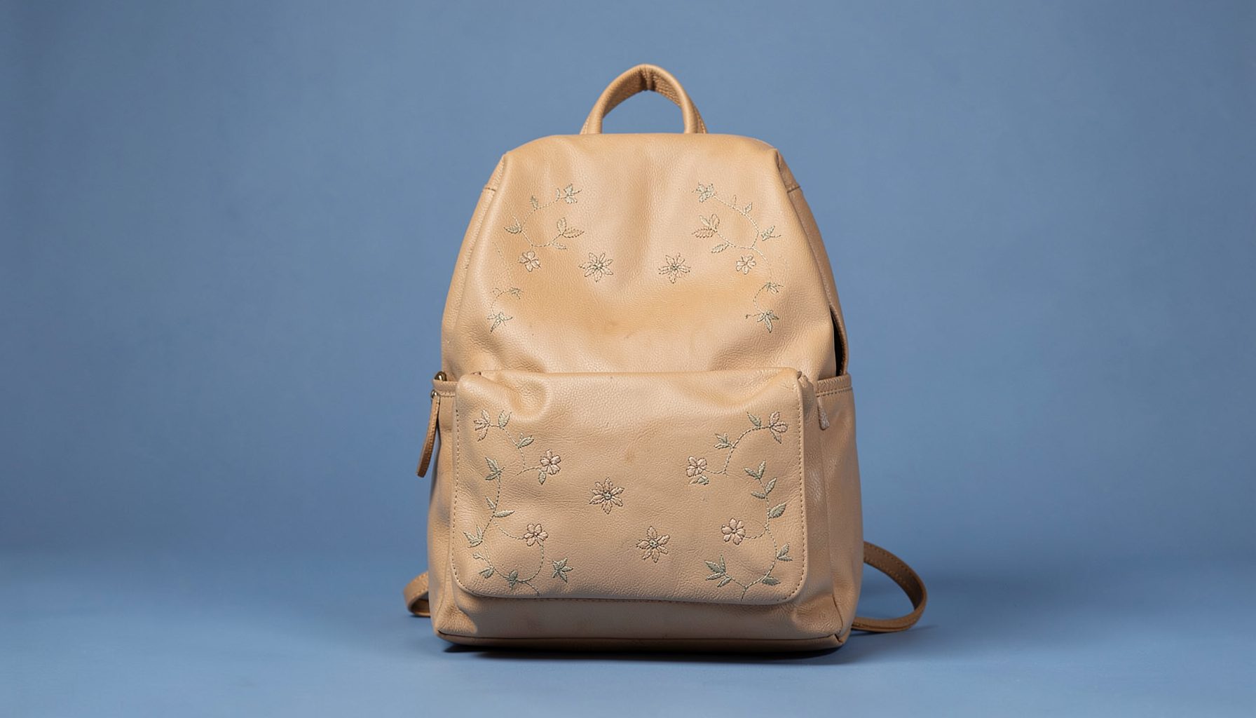 Soft navy and blue toned backdrop with a warm beige leather backpack featuring delicate embroidered floral stitching, evoking a calm vintage travel mood