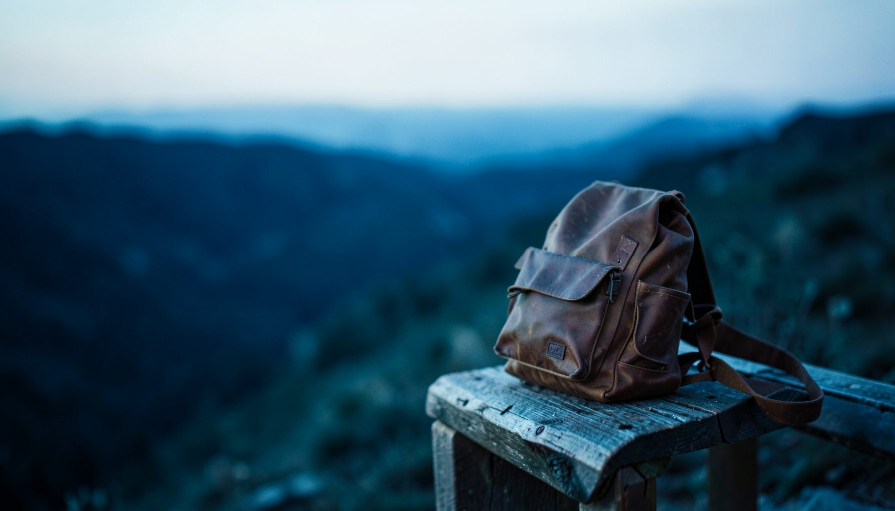 Wide travel scene in deep navy and cyan tones, a leather backpack resting on a wooden bench near a soft blurred horizon, calm and adventurous mood